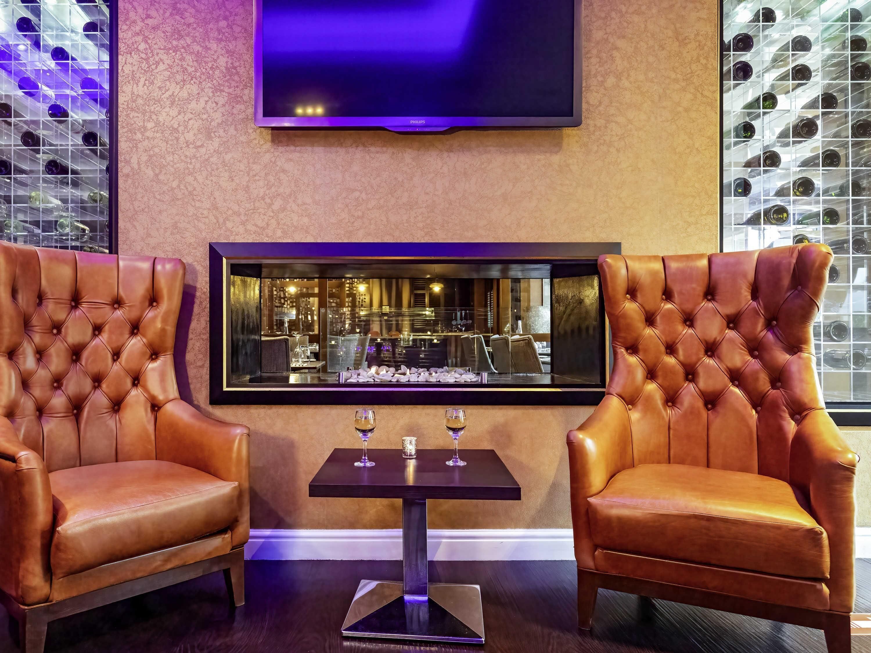 A room with two leather armchairs, a table with glasses, a flat-screen TV, and a view of a bar area.