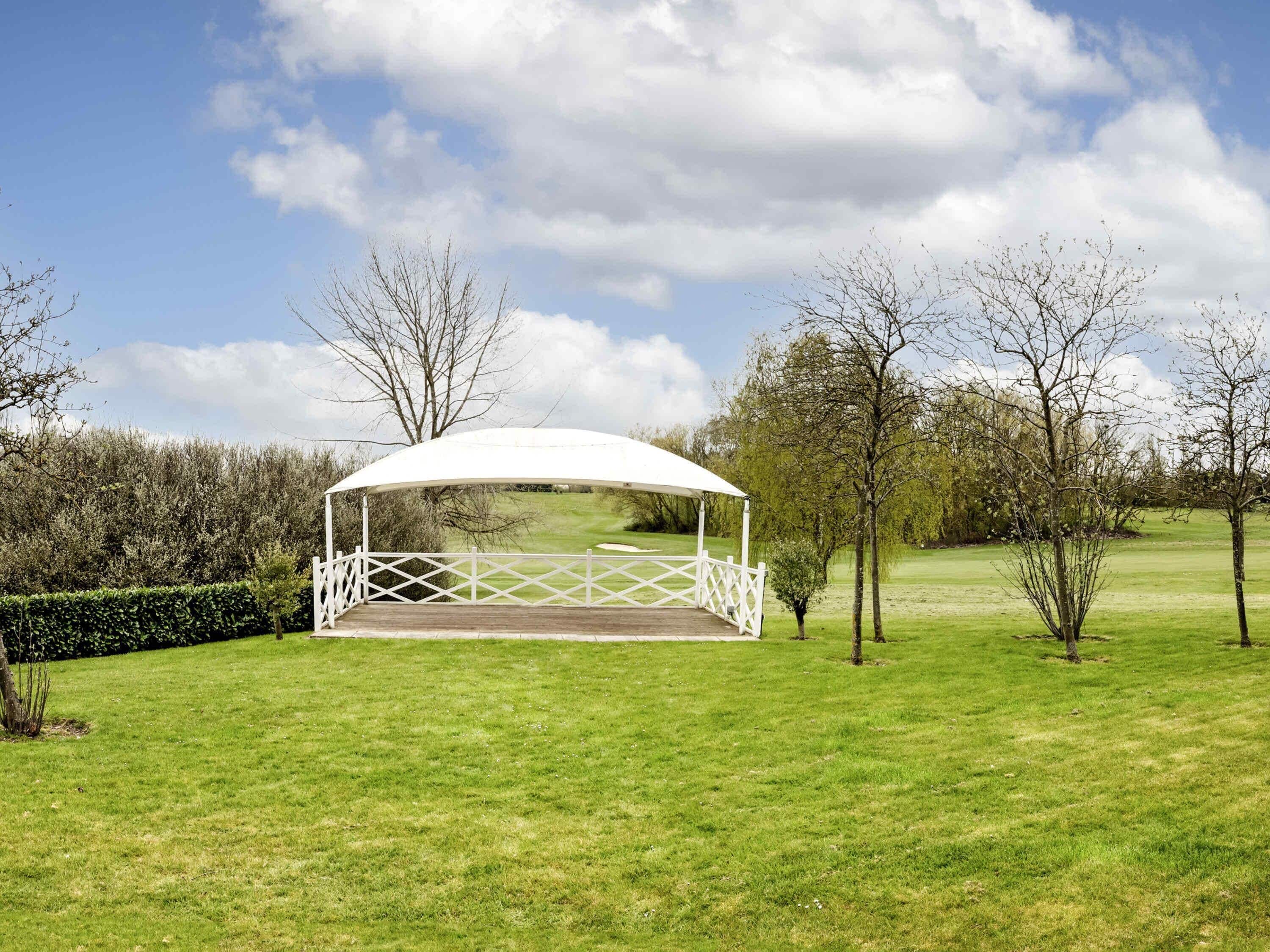 A white gazebo with a lattice base in a grassy field, surrounded by trees and hedges.