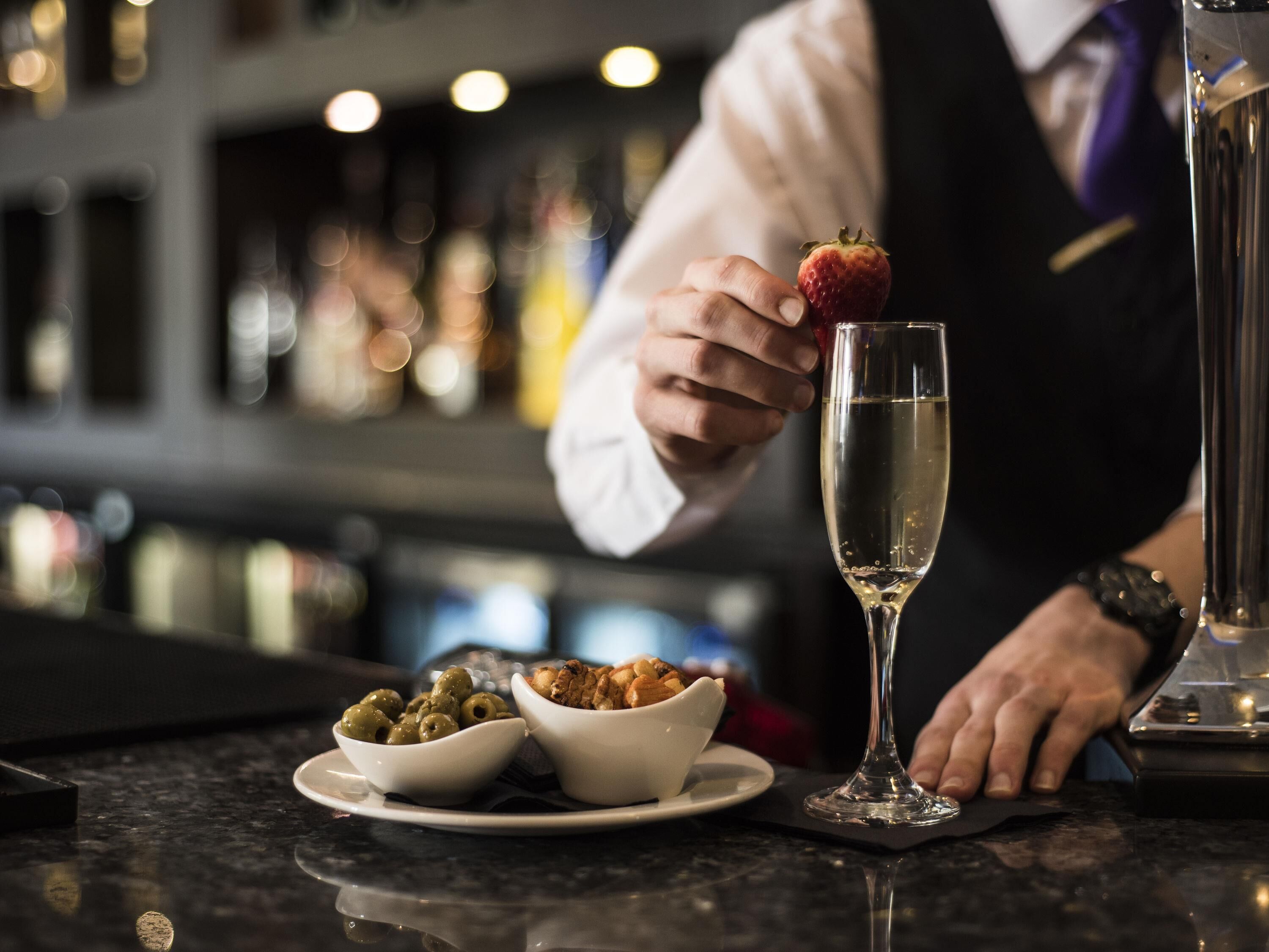 A bar setting with a bartender holding a strawberry on a glass, olives, and a bowl of food on the counter.