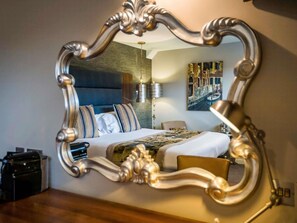 A bedroom with a large ornate mirror reflecting a bed, pillows, and a nightstand with a lamp. - Mercure Milton Keynes (Milton Keynes)