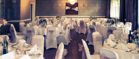 Un salón de banquetes con mesas redondas, manteles blancos y sillas con cintas blancas.