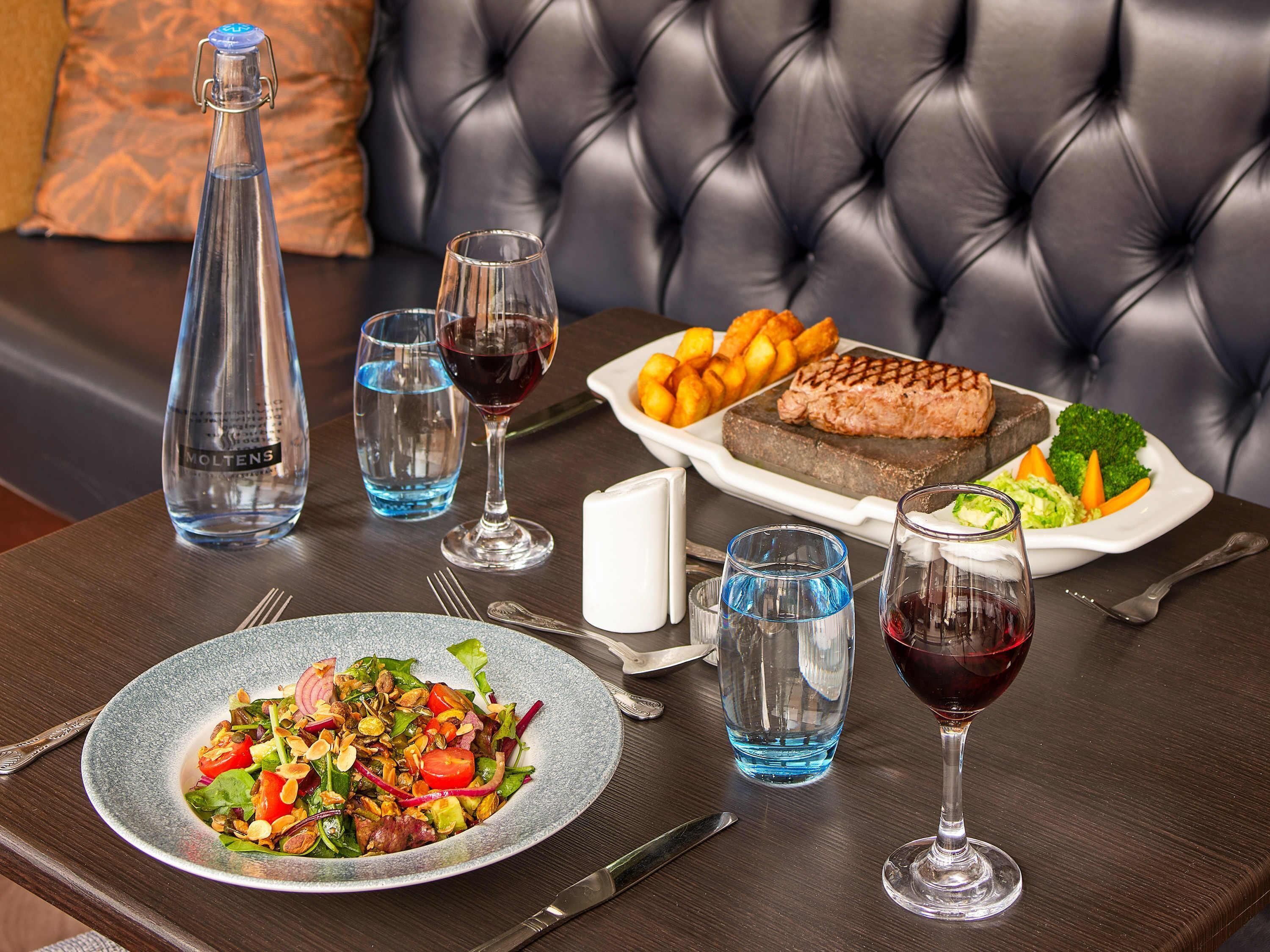 Une table à manger dressée avec une assiette de salade, un steak, des pommes de terre rôties et des légumes, ainsi que des verres à vin et une bouteille d’eau.