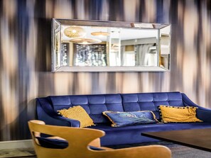 A blue sofa with two yellow pillows, a wooden chair, and a mirror on the wall. - Mercure Milton Keynes (Milton Keynes)