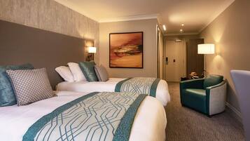 Quarto de hotel com duas camas, uma escrivaninha, uma cadeira, um quadro na parede e uma porta que dá para outro quarto.