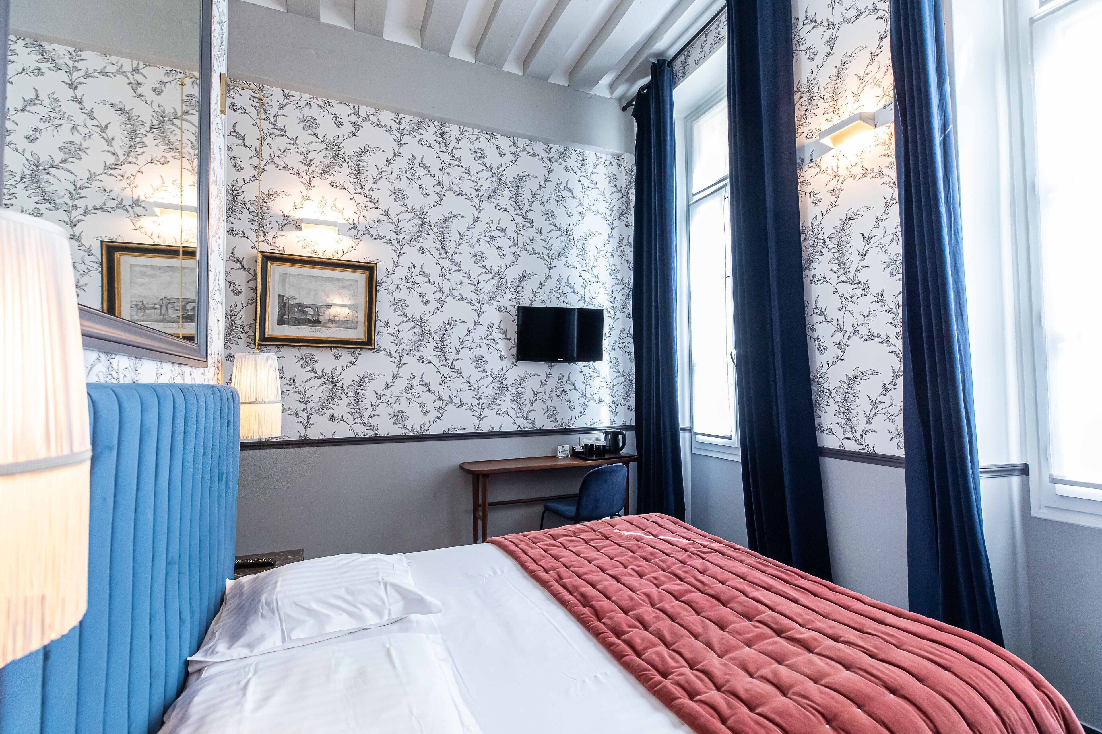 Quarto de hotel com uma cama, uma escrivaninha com cadeira, televisão e janela com cortinas.