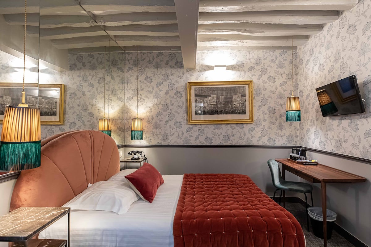A hotel room with a large bed, a desk, and a chair. There are framed pictures on the wall, a television, and a lamp.