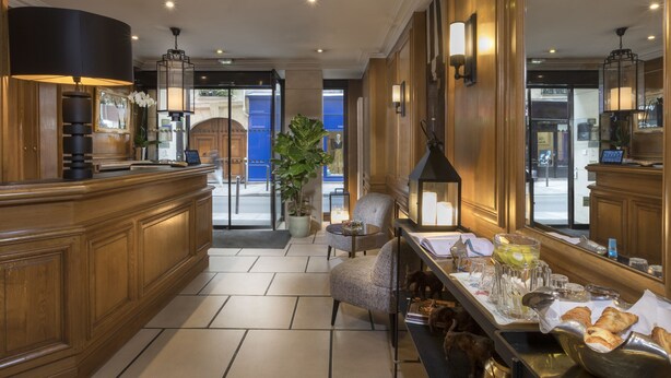 A hotel lobby with a wooden reception desk, a seating area with a table set for breakfast, and a large potted plant.