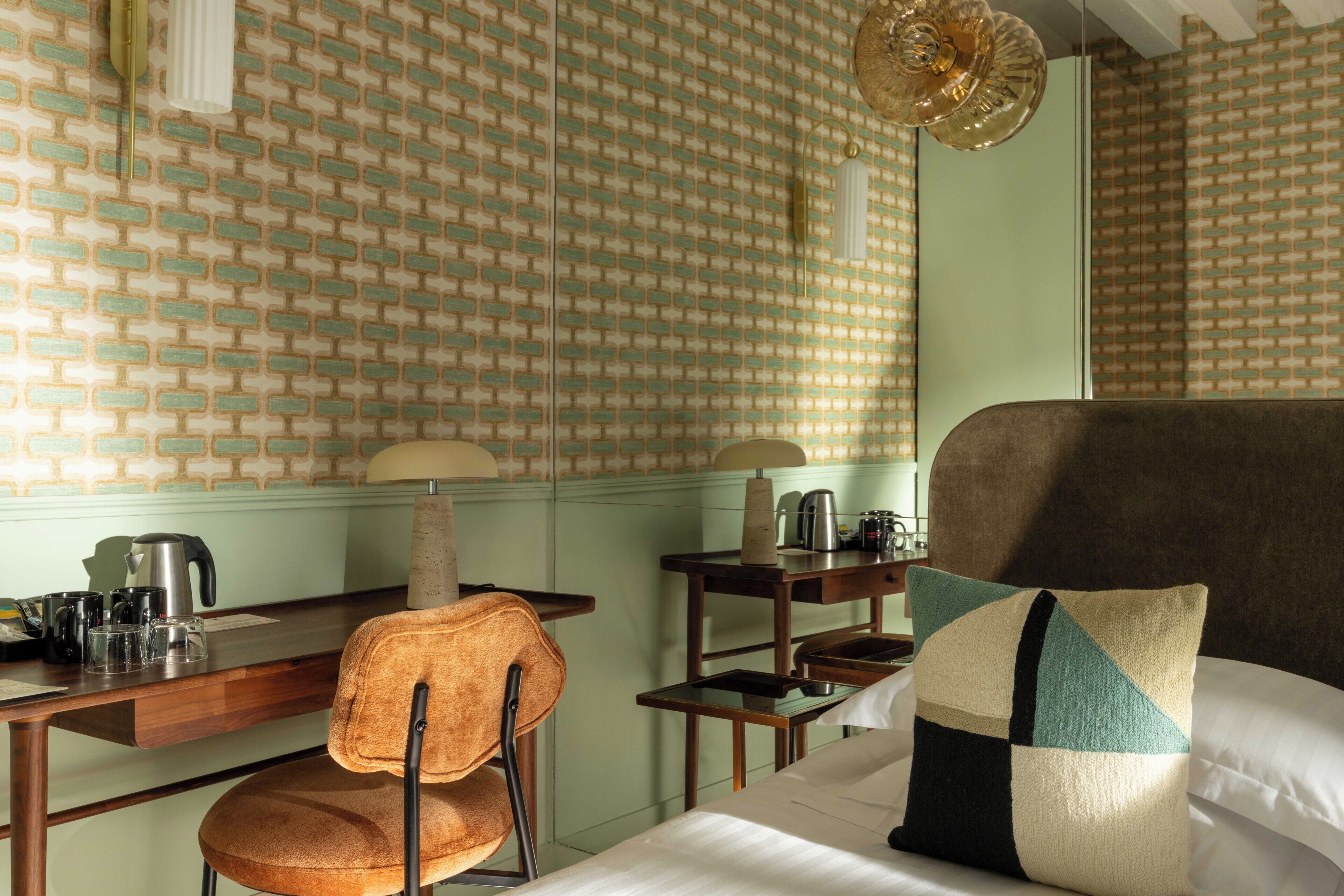 A hotel room with a brick-patterned wall, a wooden desk with a chair, a bed with a pillow, and a hanging light fixture.