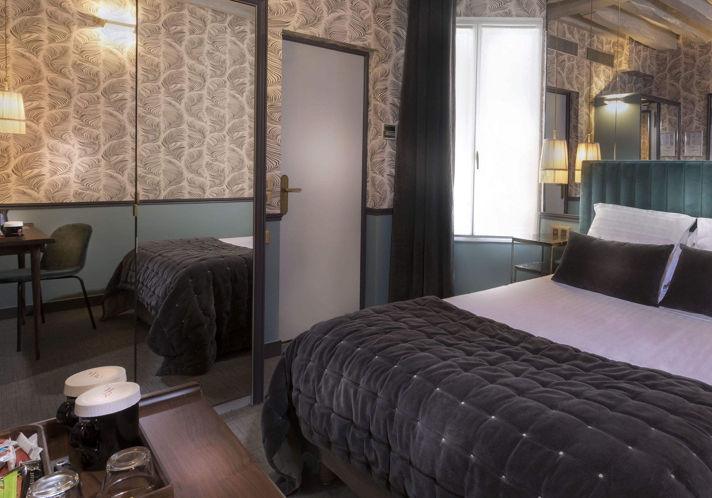 A hotel room with a bed, a desk with a chair, a bedside table, and a mirror.
