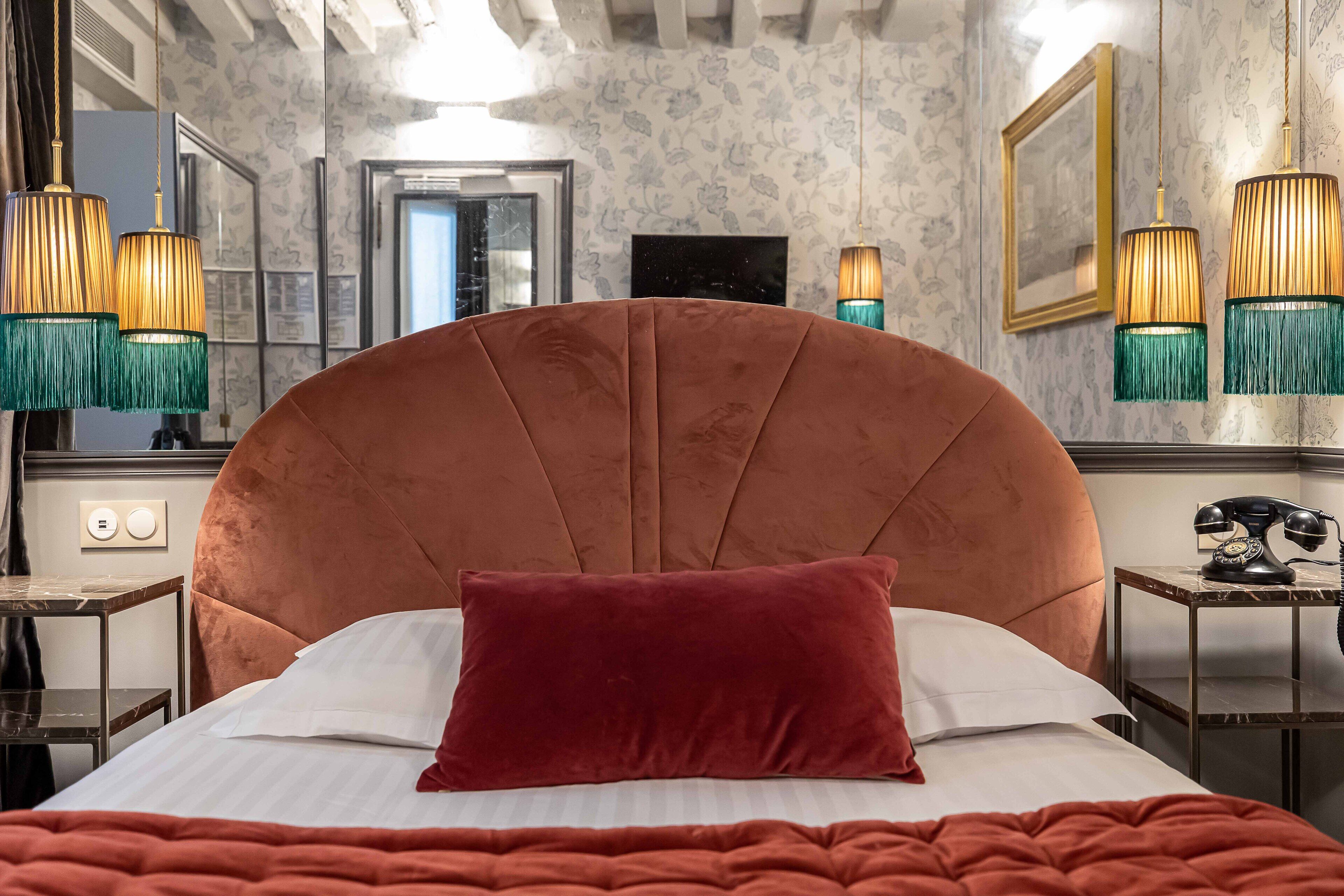A hotel room with a large, tufted headboard, a red pillow, a bed with white linens, and two bedside tables with a classic telephone and a small lamp.