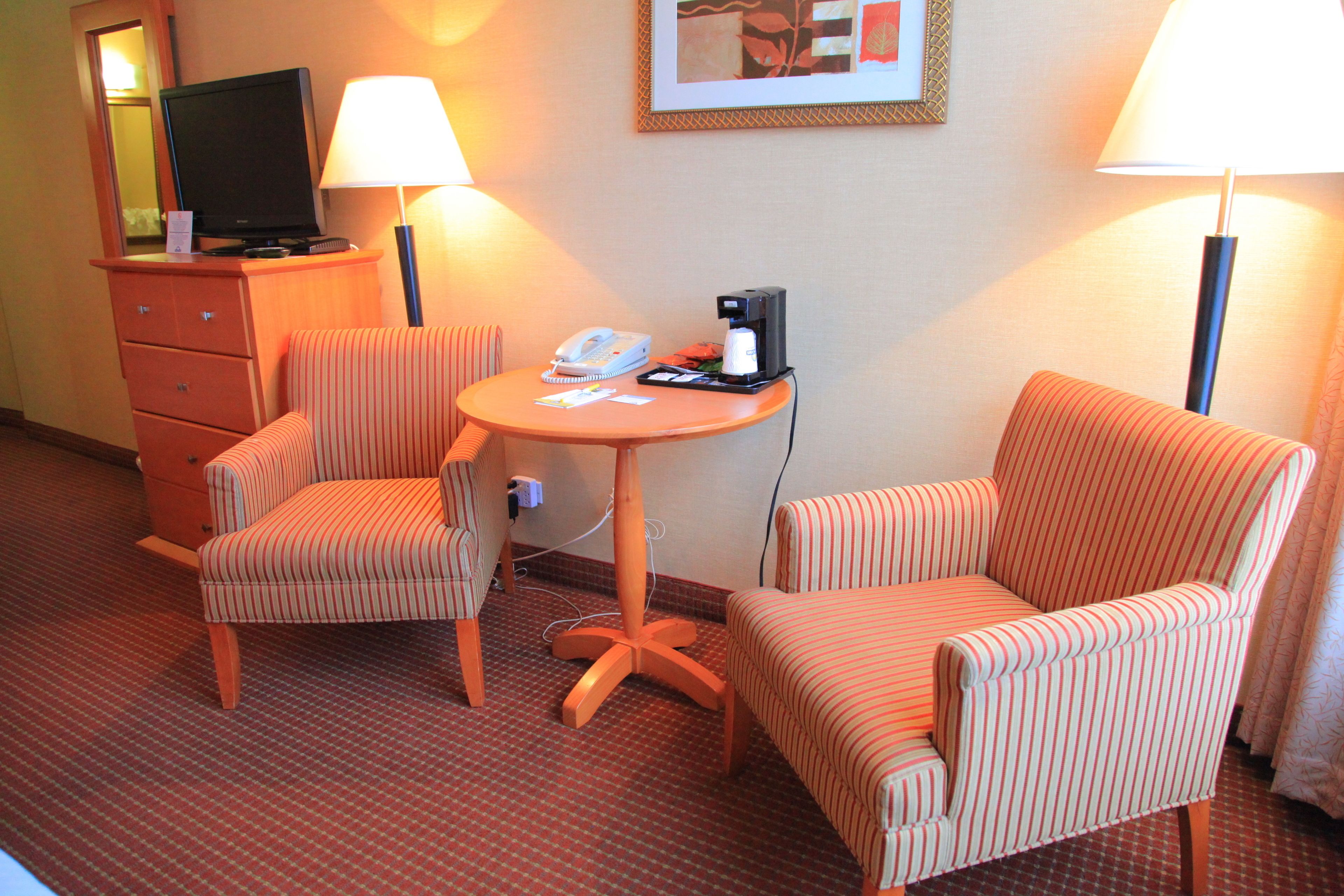 A hotel room with two striped armchairs, a small round table with a telephone and a coffee maker, and a television on a dresser.
