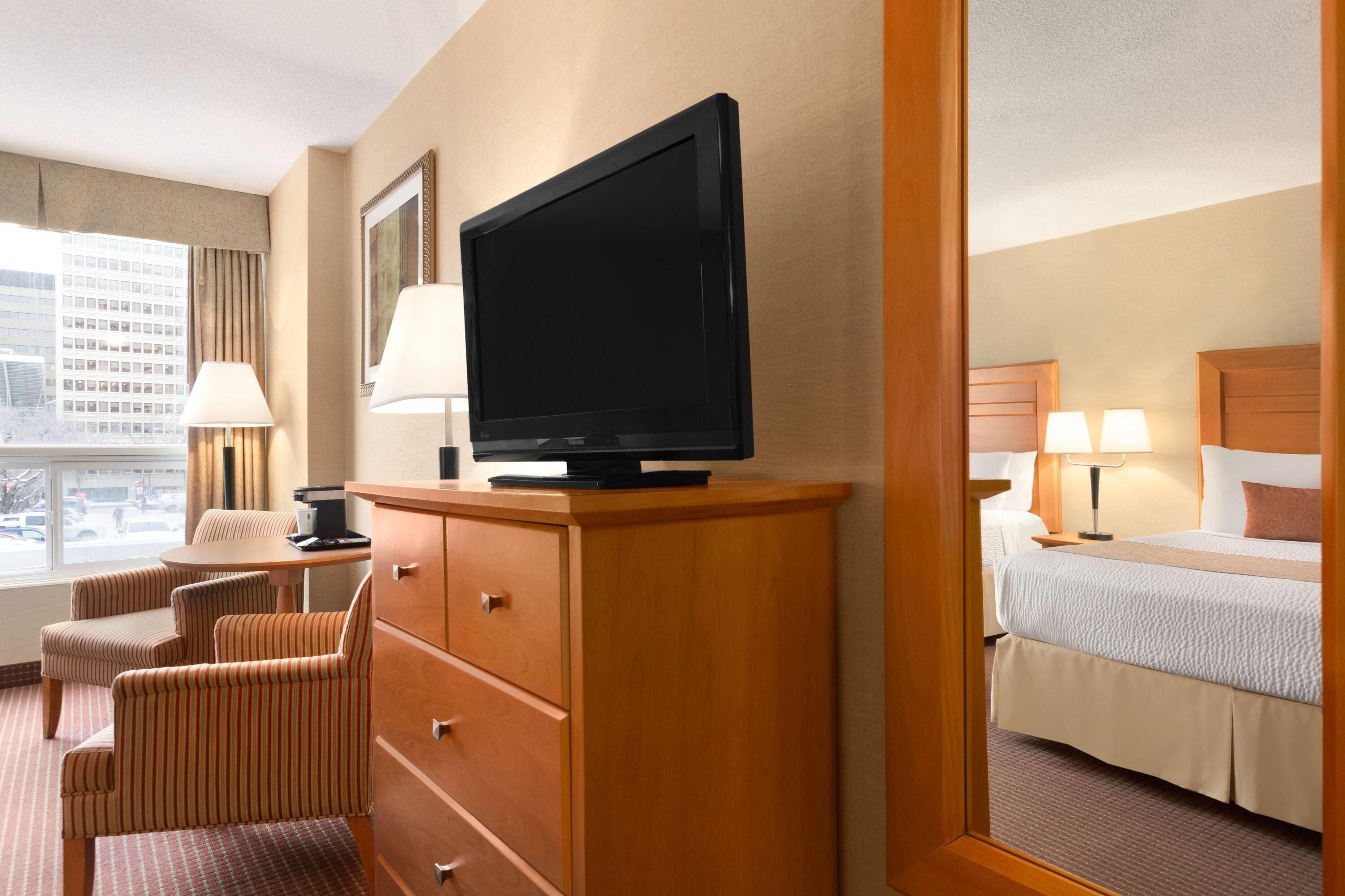 A hotel room with a television on a wooden cabinet, a desk with a lamp, striped chairs, and a bed with a headboard.
