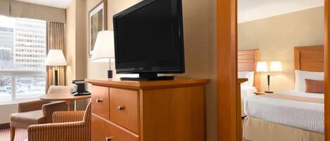 A hotel room with a television on a wooden cabinet, a desk with a lamp, striped chairs, and a bed with a headboard.