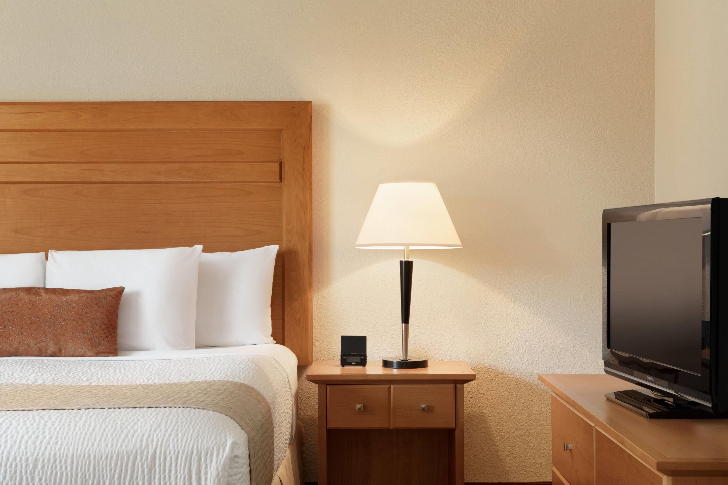 Ein Hotelzimmer mit einem hölzernen Kopfteil, einem Bett mit weißen und braunen Kissen, einer Nachttischlampe, einem Fernseher auf einer Holzkommode und einer kleinen Schubladenkommode.