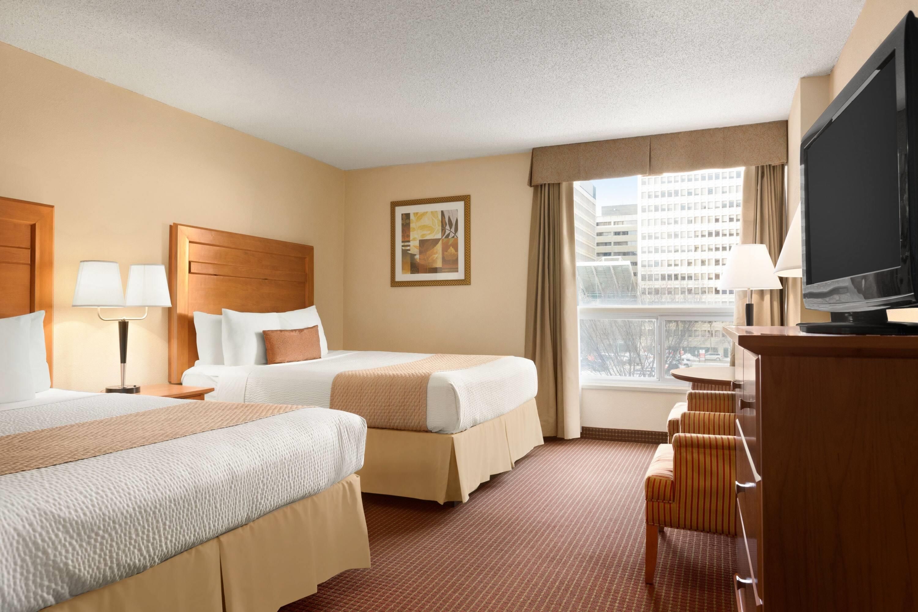 A hotel room with two beds, a desk, a television, and a large window with a city view.