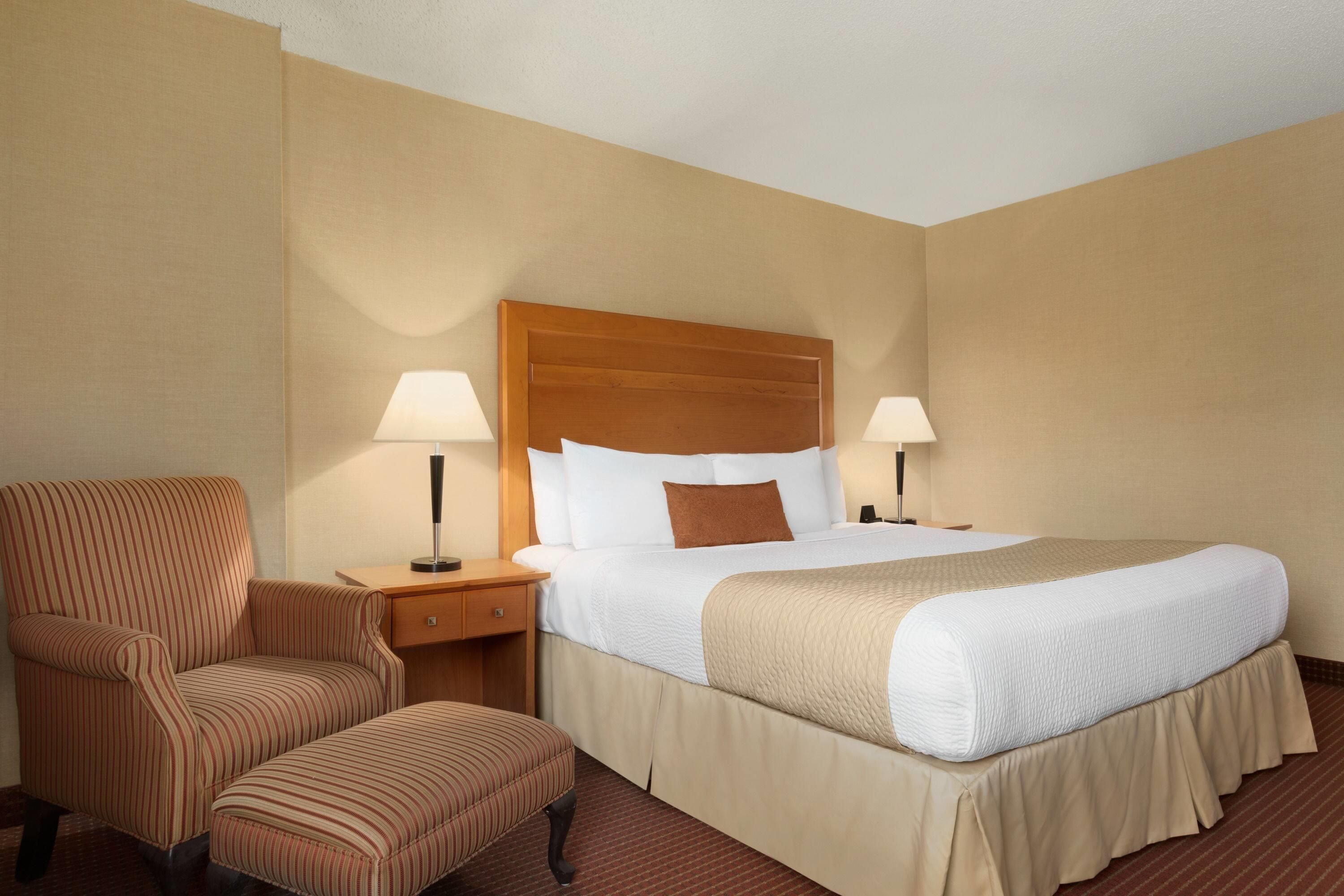 A hotel room with a bed, two bedside lamps, a striped armchair, and a small table.