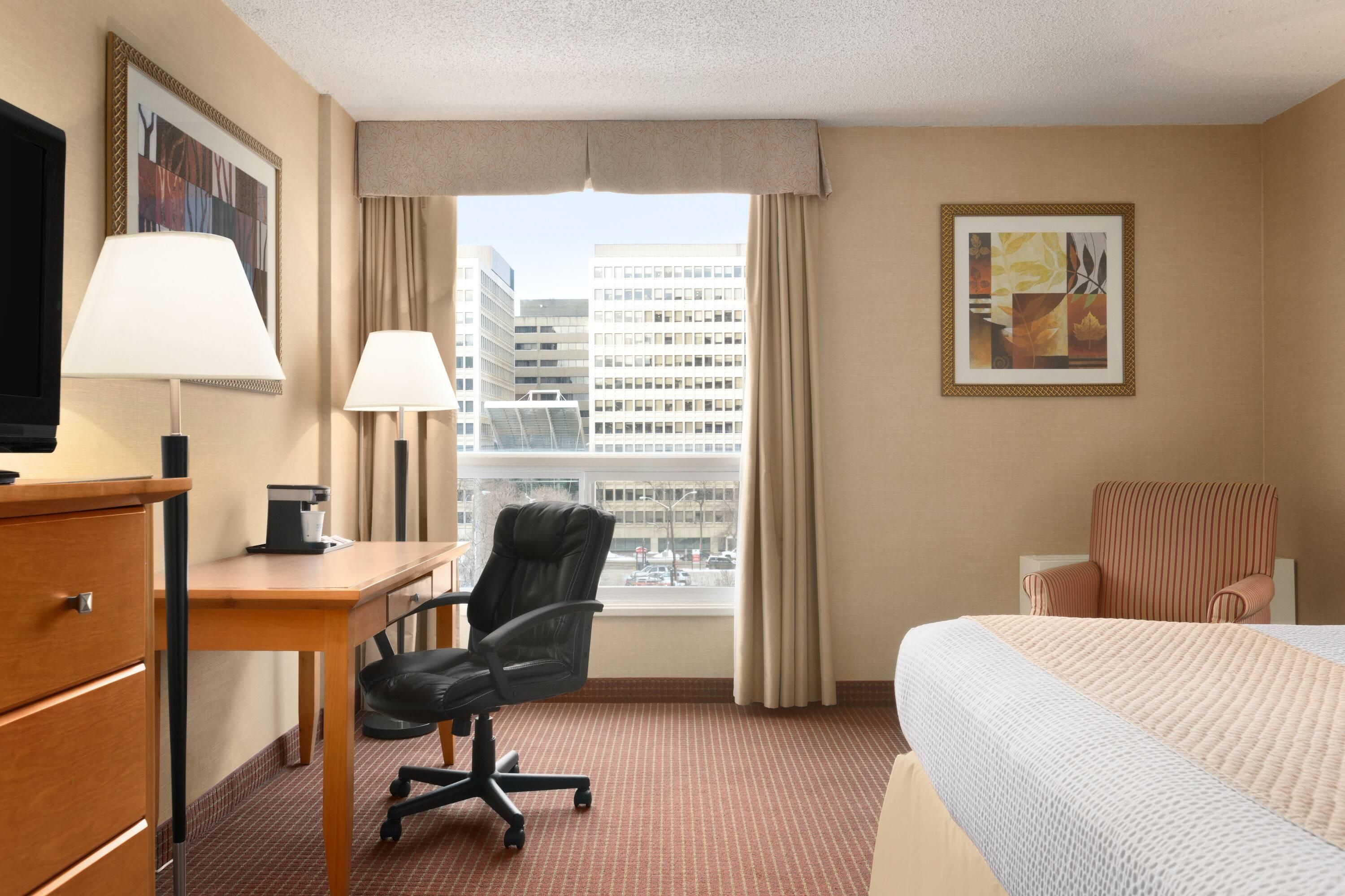 Une chambre d’hôtel équipée d’un lit, d’un bureau, d’une chaise et d’une grande fenêtre donnant sur la ville.