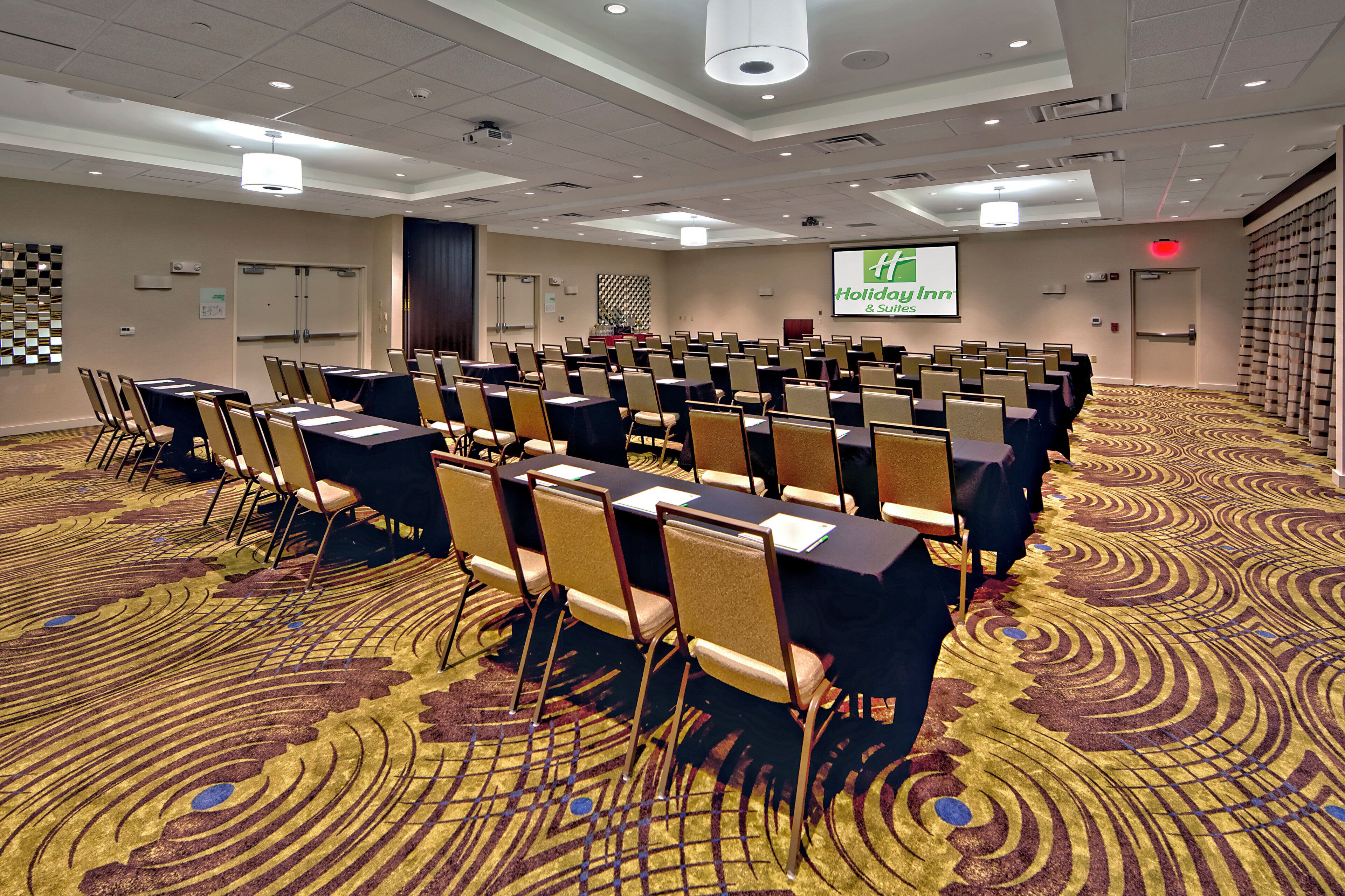 A conference room with rows of tables and chairs, a projector screen, and a carpeted floor.