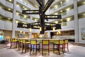 Un hall d’hôtel spacieux doté d’un bar central, de plusieurs espaces aménagés pour s’asseoir et d’un haut plafond laissant entrer la lumière naturelle.