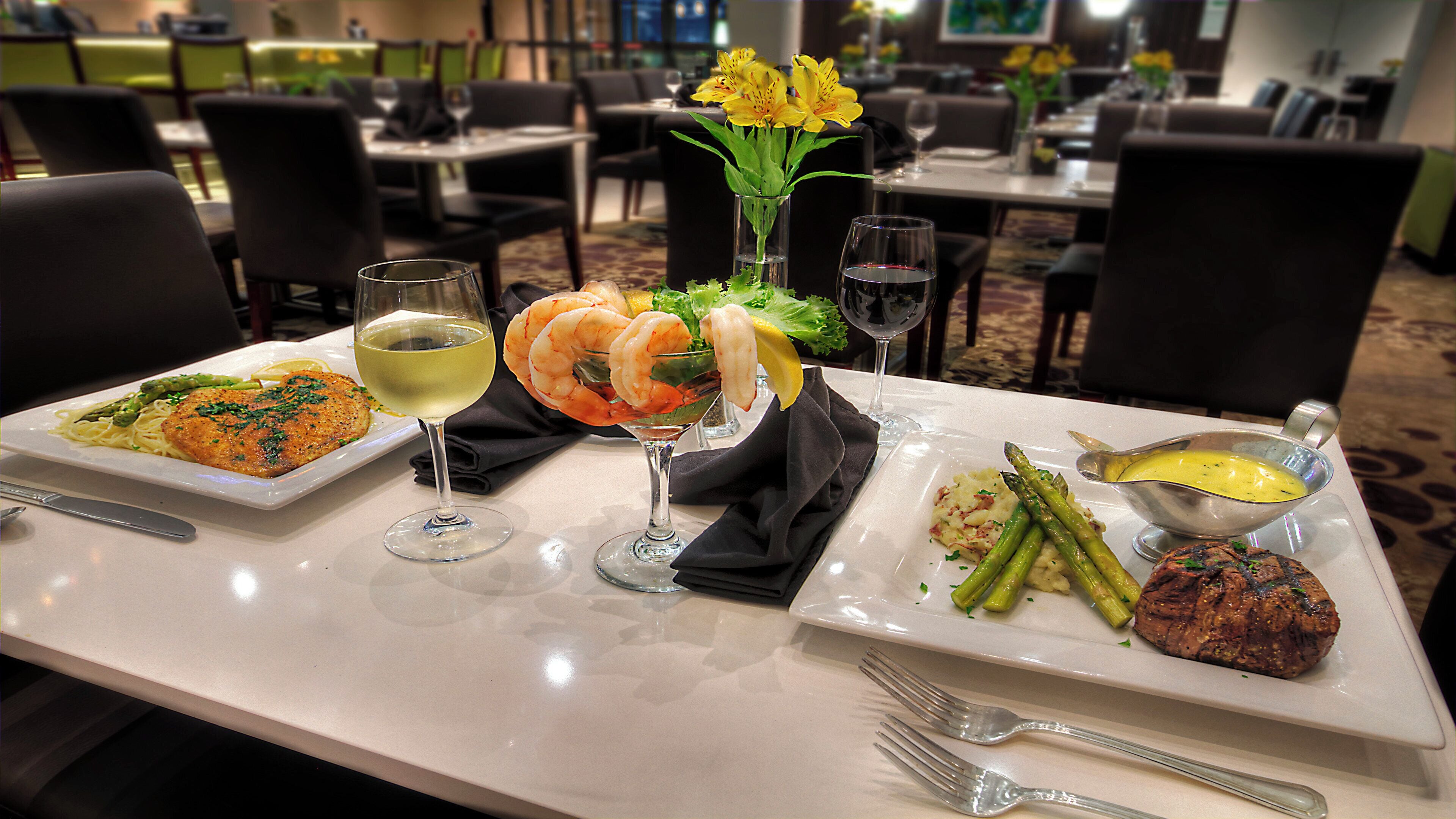 Una mesa de comedor con diversos platos, incluyendo mariscos, verduras y un filete, junto con copas de vino y cubiertos.