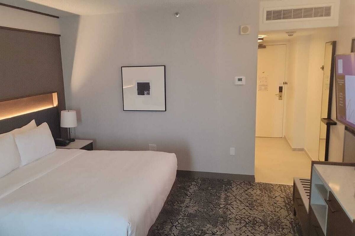 A hotel room with a large bed, a bedside table with a lamp, a framed picture on the wall, and a carpeted floor.