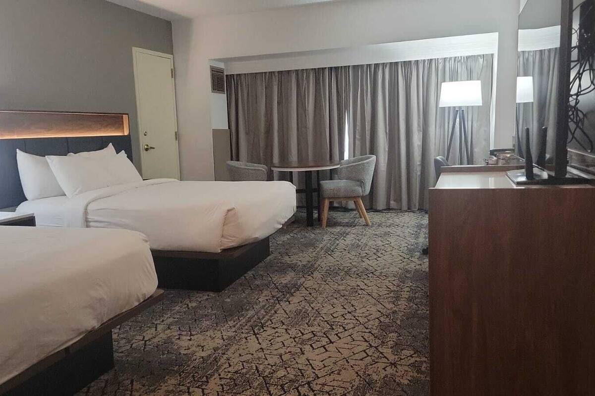 A hotel room with two beds, a desk, a chair, and a large window with curtains.