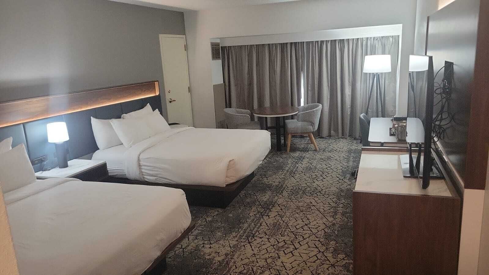Habitación de hotel con dos camas, una mesita, sillas, televisor y ventana con cortinas.