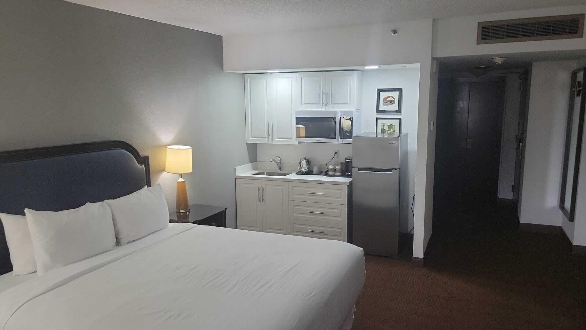 A hotel room with a bed, a nightstand, a lamp, a kitchenette with a microwave, and a refrigerator.