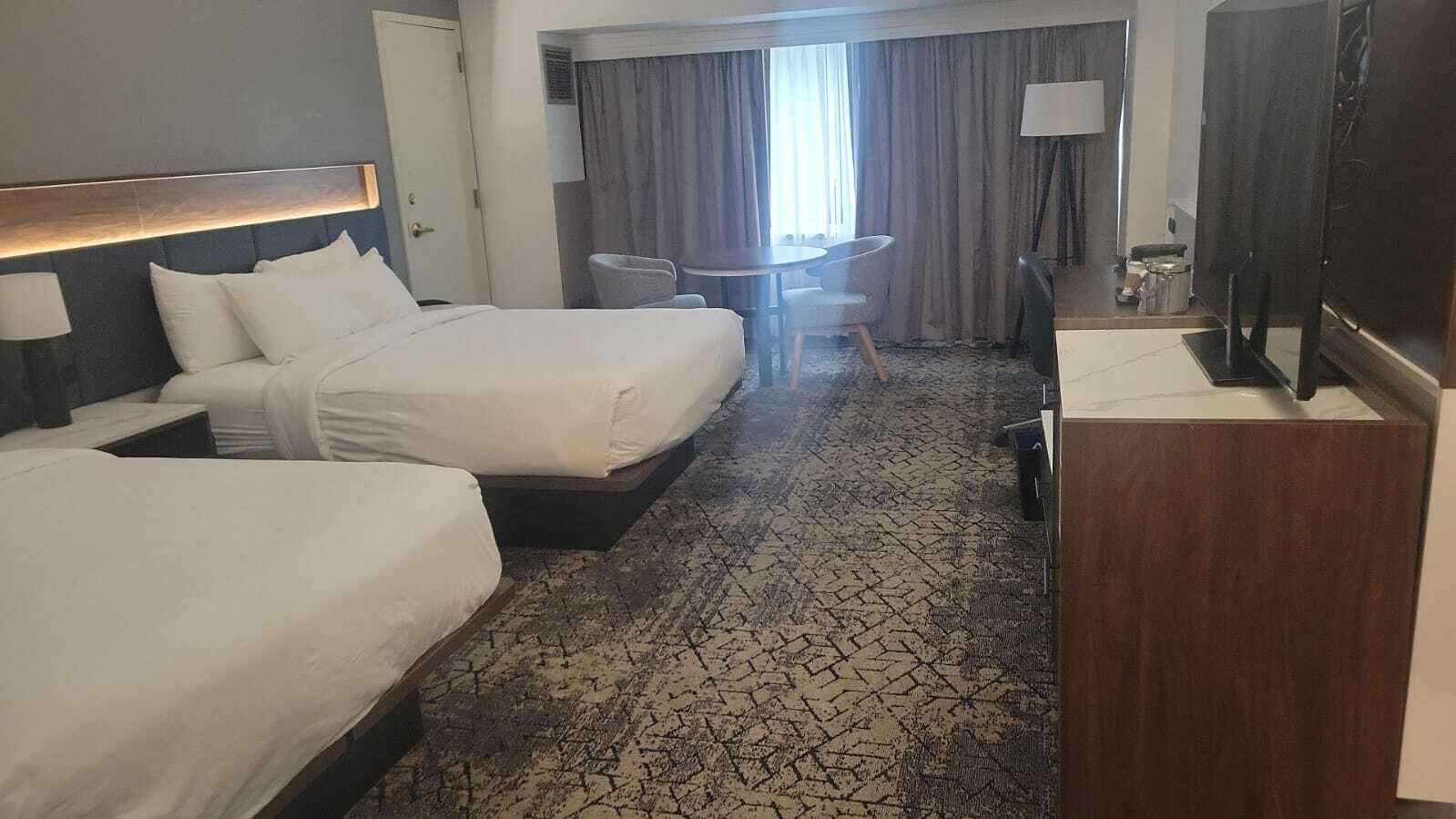 A hotel room with two beds, a desk, a chair, a TV, and a window with curtains.