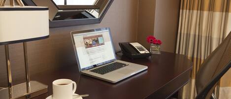 A hotel room desk with a laptop, a cup of coffee, a phone, and a vase with flowers.