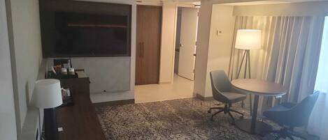 Habitación de hotel con escritorio, silla, mesa, lámpara y un ventanal con cortinas.