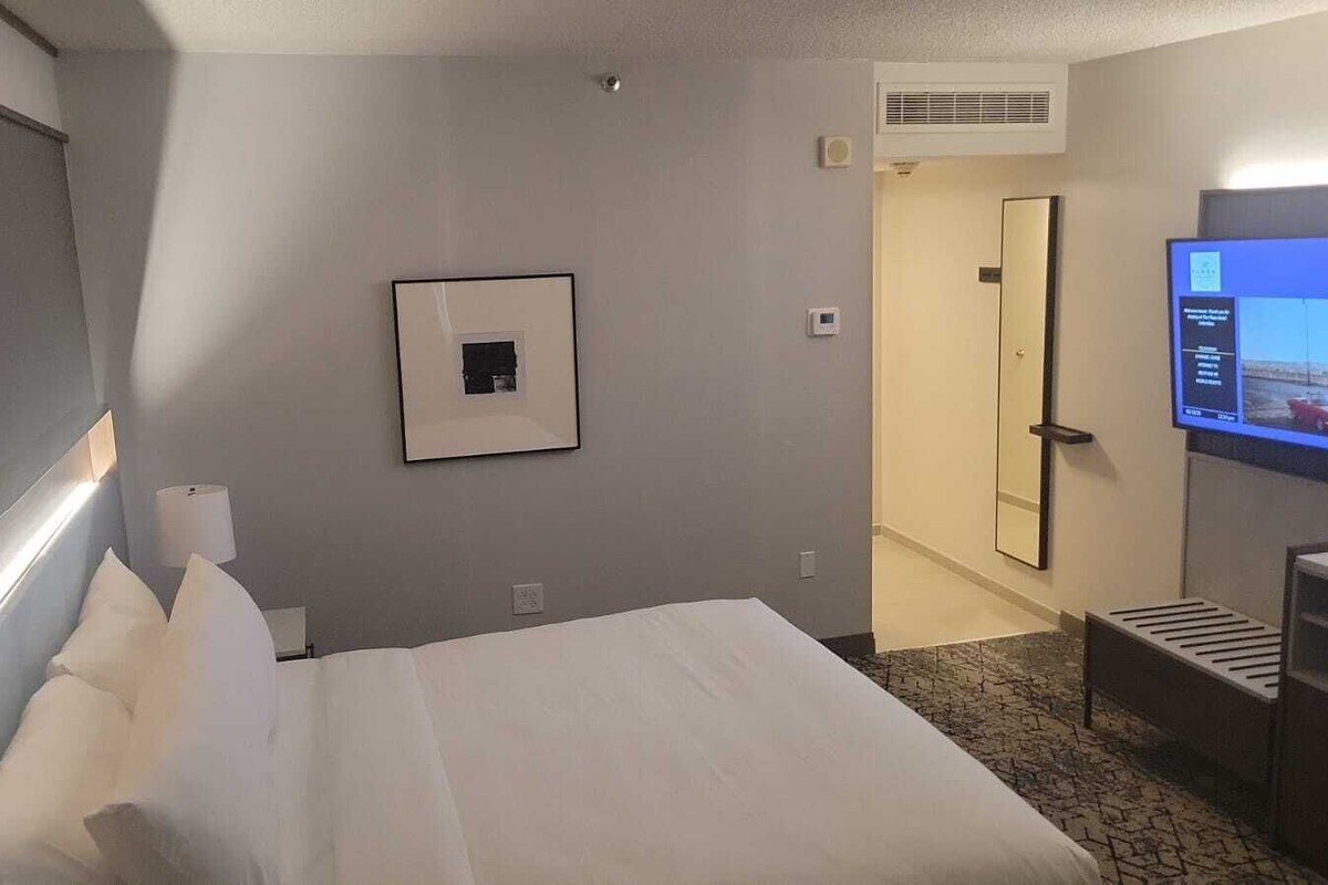Une chambre d’hôtel avec un lit, une télévision, un tableau encadré au mur et une porte donnant sur une autre pièce.