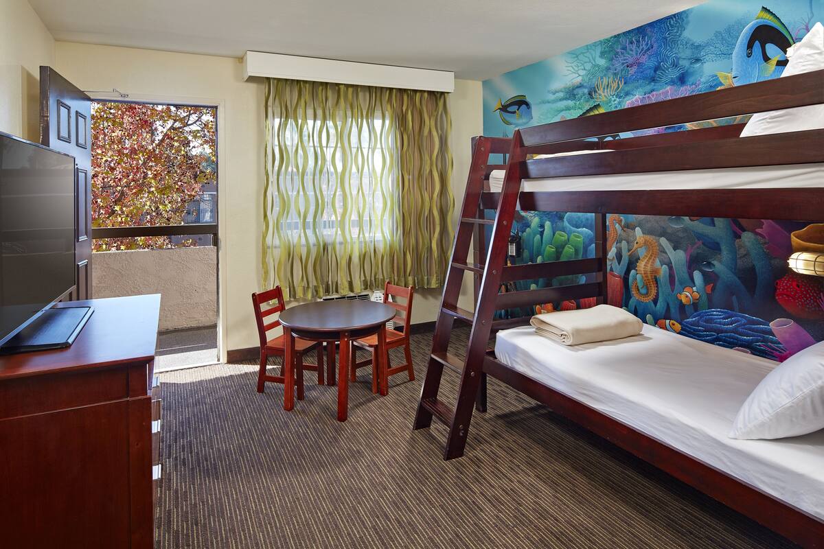 A hotel room with a bunk bed, a desk with a laptop, a dining table with chairs, and a large mural of an underwater scene.