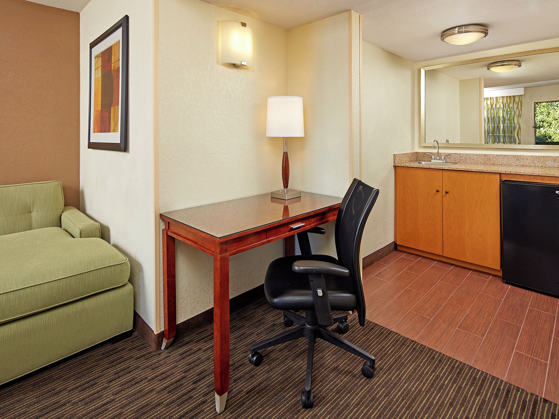A hotel room with a green sofa, a wooden desk, a black office chair, a lamp, a kitchen area with a sink, and a refrigerator.