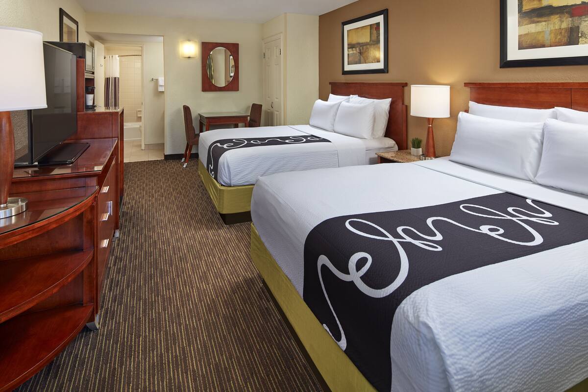 A hotel room with two beds, a desk with a television, a chair, and a small table with a lamp.