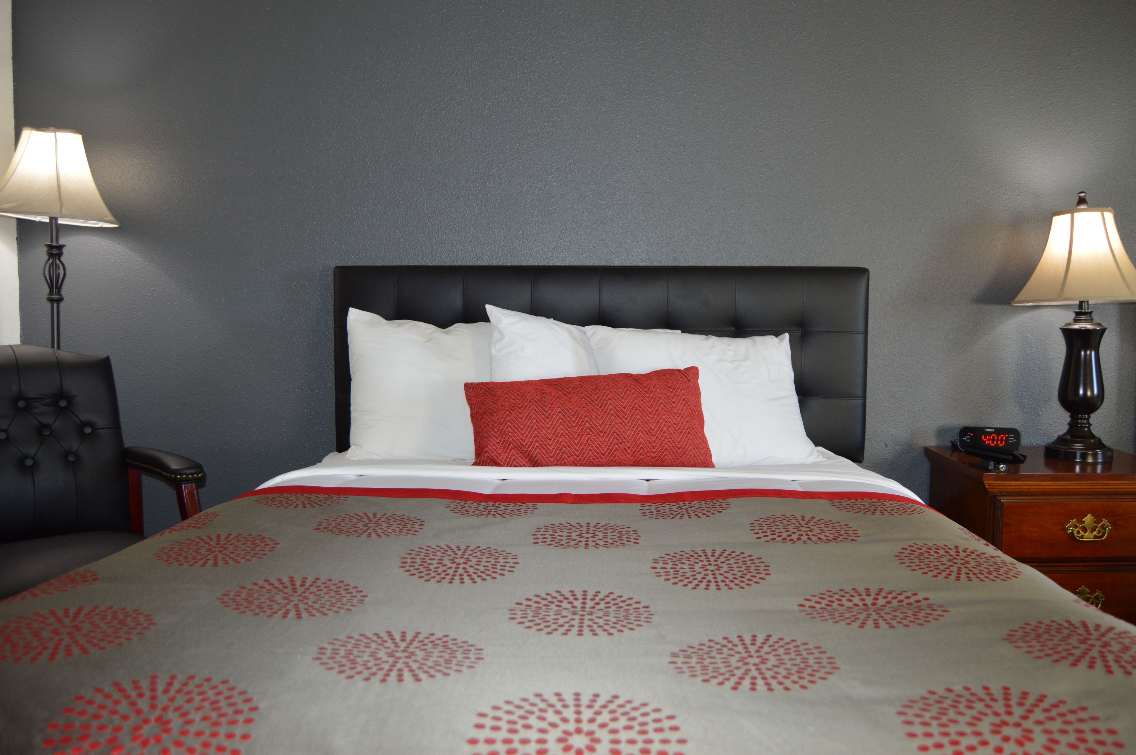 Una cama bien hecha con una colcha blanca y roja estampada, un cabecero tapizado de color negro y dos mesitas de noche con lámparas.