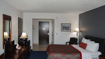 Habitación de hotel con cama, escritorio con lámpara, silla, televisor y puerta que da a otra habitación.