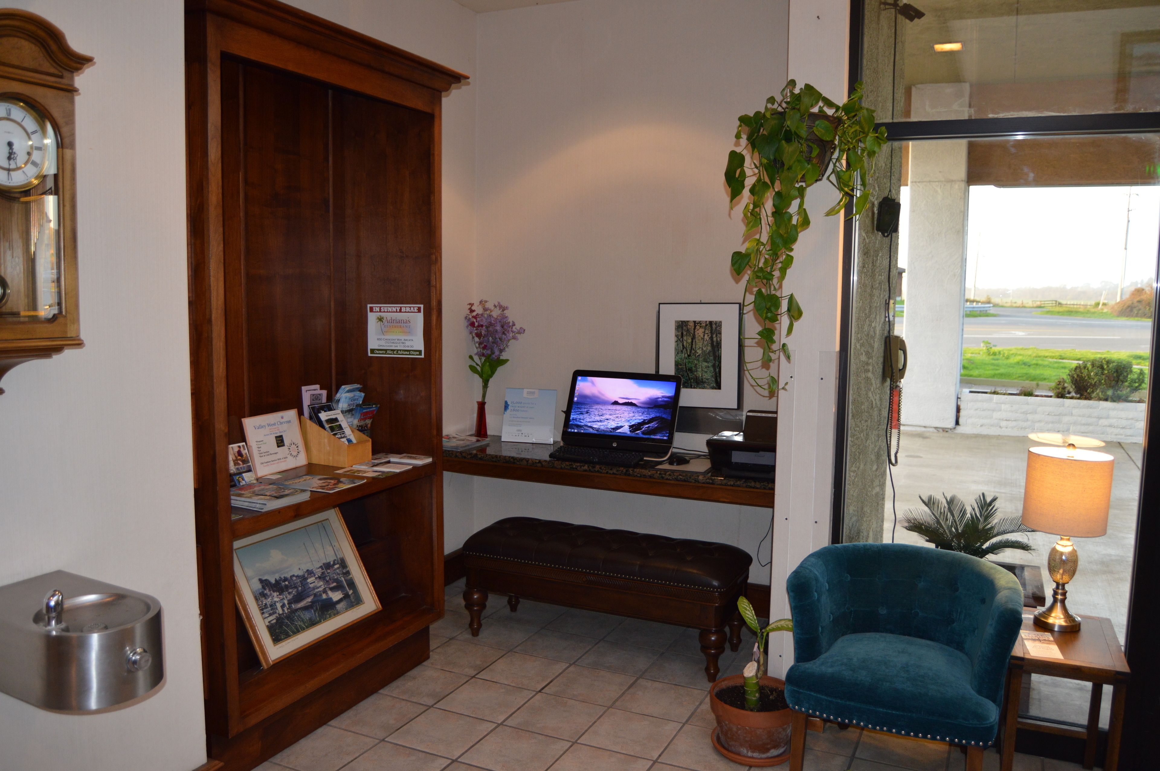 Un área de recepción con un escritorio de madera, una computadora portátil, un cuadro enmarcado, un banco, una planta en maceta y un reloj de pared.
