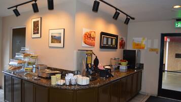 Zona de desayuno de hotel con barra de buffet, que incluye diversas opciones de comida y una máquina de café.