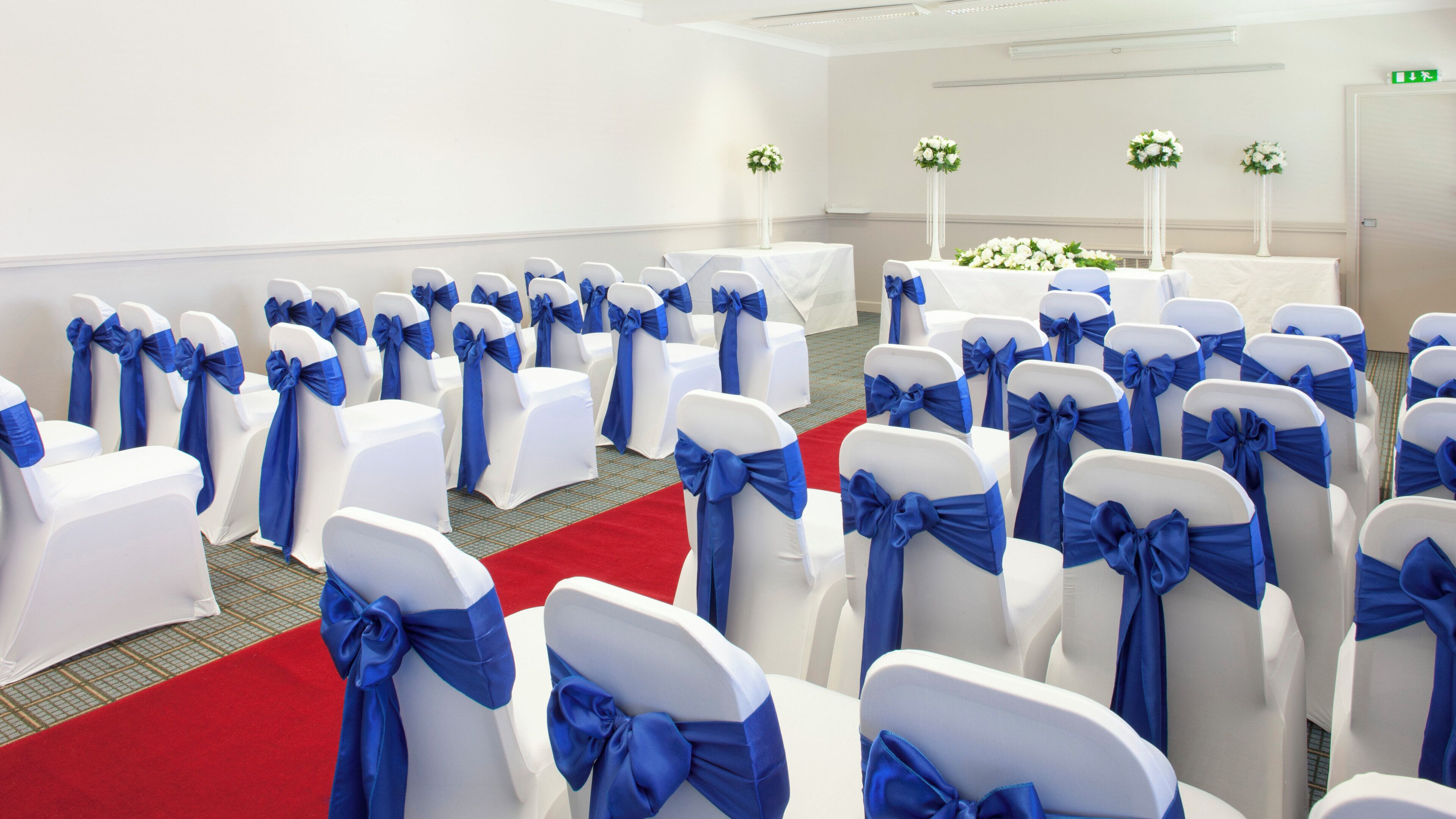 A conference room with rows of white chairs with blue sashes, a red carpet, and floral centerpieces on tables.