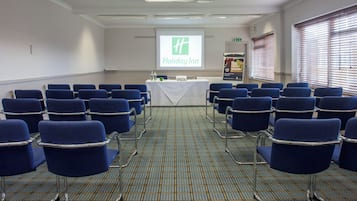 Uma sala de conferências com fileiras de cadeiras azuis, uma mesa branca e o logotipo da Holiday Inn na tela.