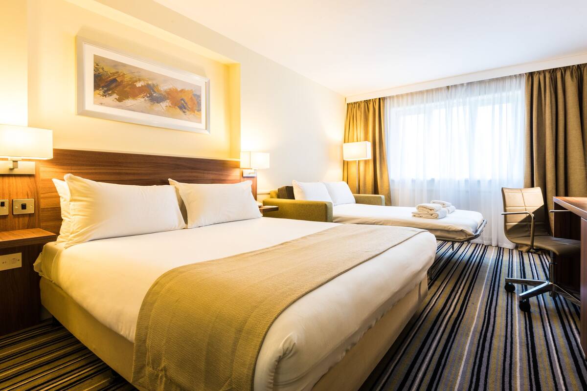 A hotel room with two beds, a desk, a chair, and a large window with curtains.