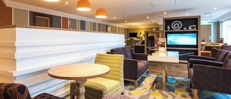 A modern hotel lobby with a reception desk, seating area, and a television screen.