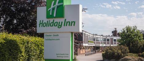 Señal de Holiday Inn con logotipo verde, un área de recepción y un edificio al fondo.