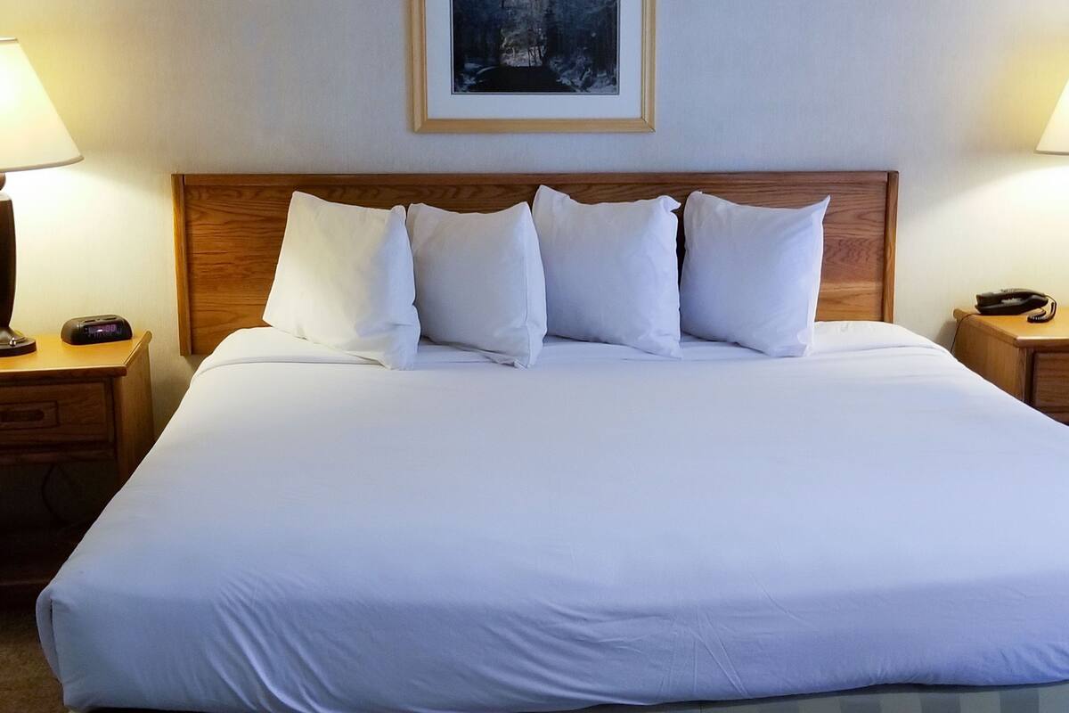 Una cama bien hecha con ropa blanca y un cabecero de madera. Hay dos mesitas de noche con lámparas y un cuadro enmarcado en la pared sobre la cama.