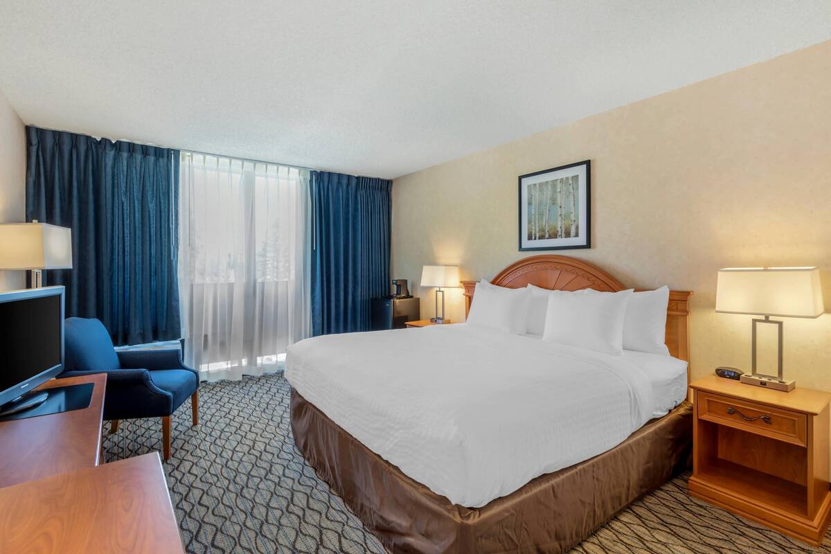 A hotel room with a large bed, a desk with a TV, a chair, two bedside tables with lamps, and a window with curtains.