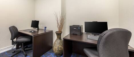 A hotel room with a desk, two monitors, a laptop, a chair, a vase with branches, and a carpet with a blue pattern.