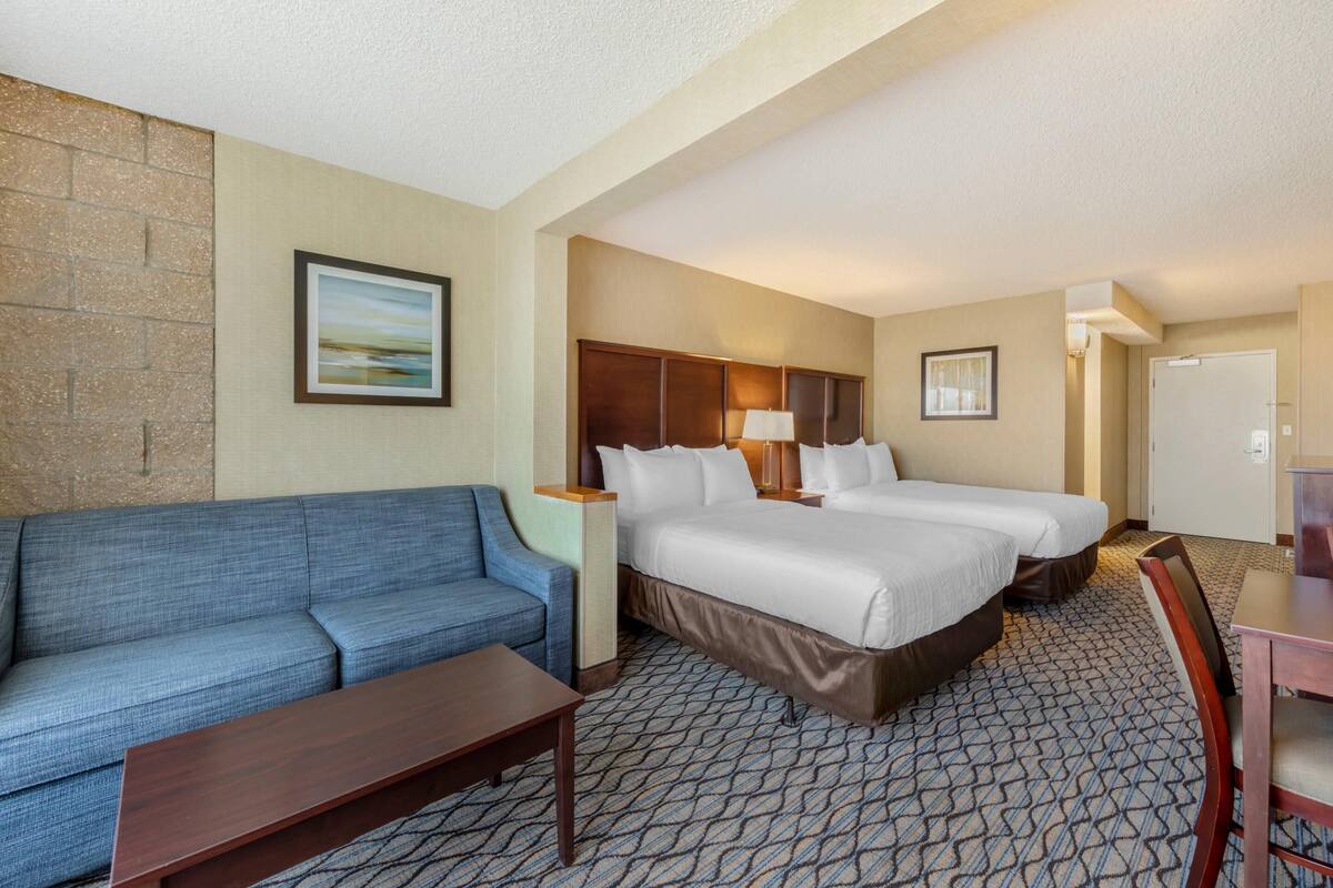 A hotel room with a blue sofa, two beds, a wooden nightstand, a lamp, and a patterned carpet.