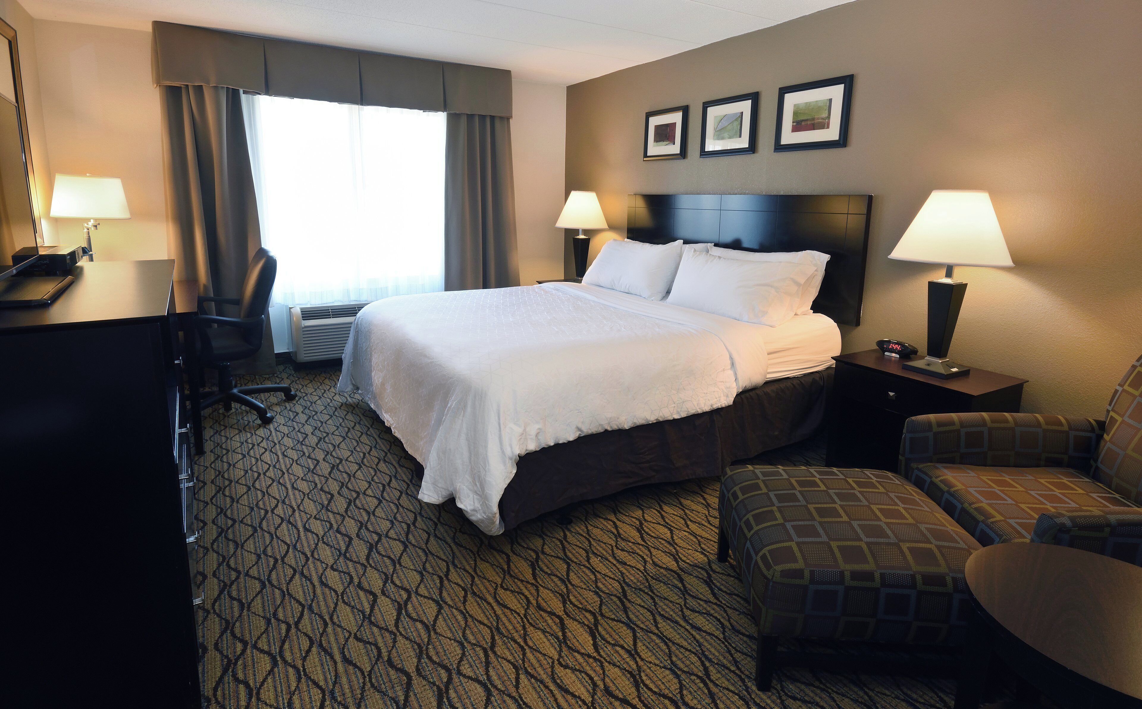 A hotel room with a large bed, two bedside lamps, a desk with a computer, a chair, and a patterned carpet.