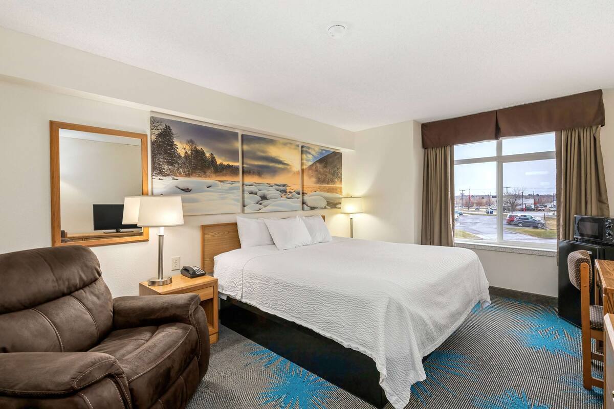 A hotel room with a large bed, a brown leather chair, a desk with a lamp, a television, and a window with curtains.